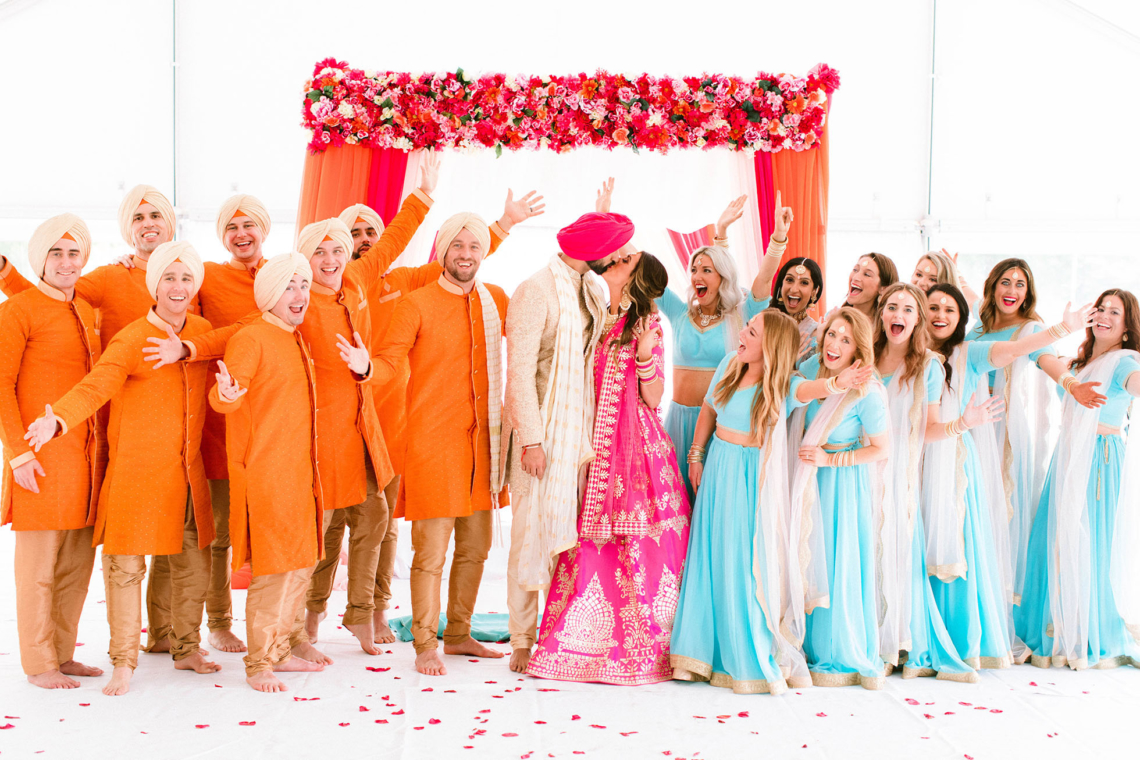 Bride and Groom kiss with wedding party surrounding them all wearing traditional Indian wedding clothes
