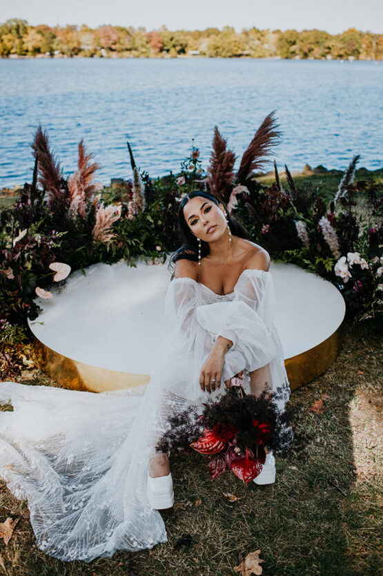 bride poses sitting on round platform at end of ceremony aisle with lake in background