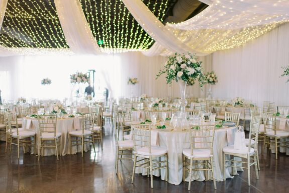 Wedding Reception setup inside Lakeview Event Center with canopy of string lights over classic white decor with modern acrylic flower stands with romantic rose arrangements