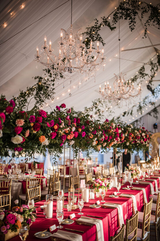 tented wedding reception with elevated bright pink florals, and greenery, pink linens, hanging string lights, and chandeliers.