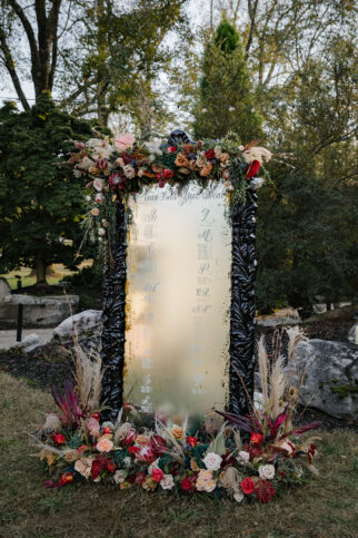 Wedding seating chart on tall black-framed floor mirror adorned with fall-colored flowers and dried pampas grass