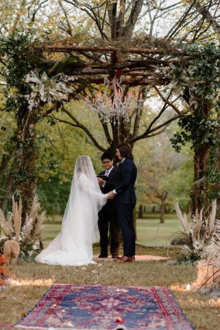 Gray and Billy hold hands at the altar during their ceremony beneath an arch created with tree branches and greenery