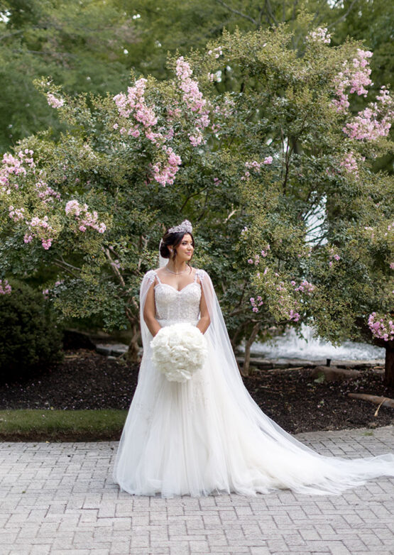 Bride in Lace Gown with Corset Bodice, Matching Bridal Cape and Bejeweled Tieara with Classic Bun Updo and Face-Framing Curls | Full Round White bouquet