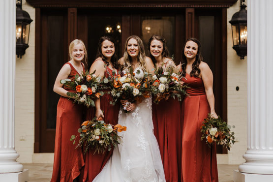 boho bride and bridesmaid in rust colored dressed and fall themed florals pose infront of the estate