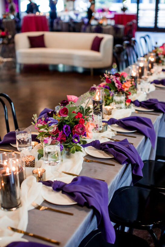 Wedding reception table setup with purple napkins, bright colorful arrangements, and moody candles