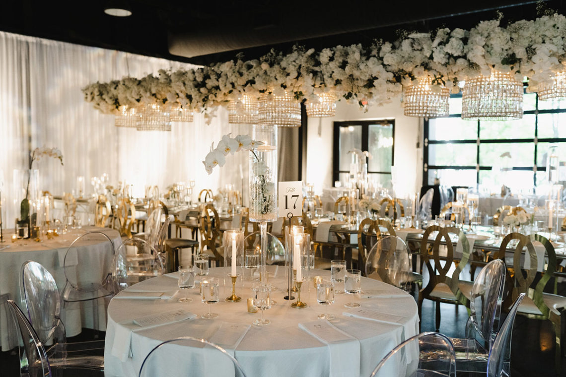 Wedding Reception with Lush Hanging White Floral Installation, Crystal Chandeliers, Ghost Chairs, and Mirrored Kings Tables