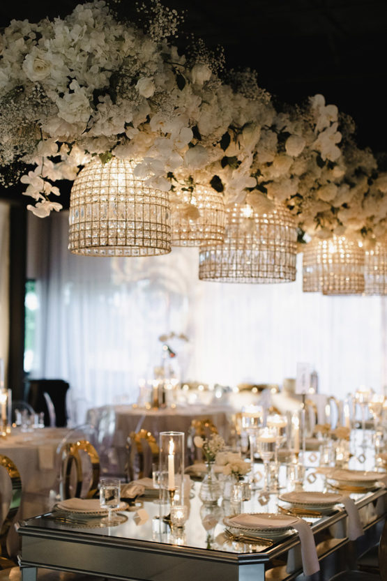 Wedding Reception with Lush Hanging White Floral Installation, Crystal Chandeliers, Ghost Chairs, and Mirrored Kings Tables