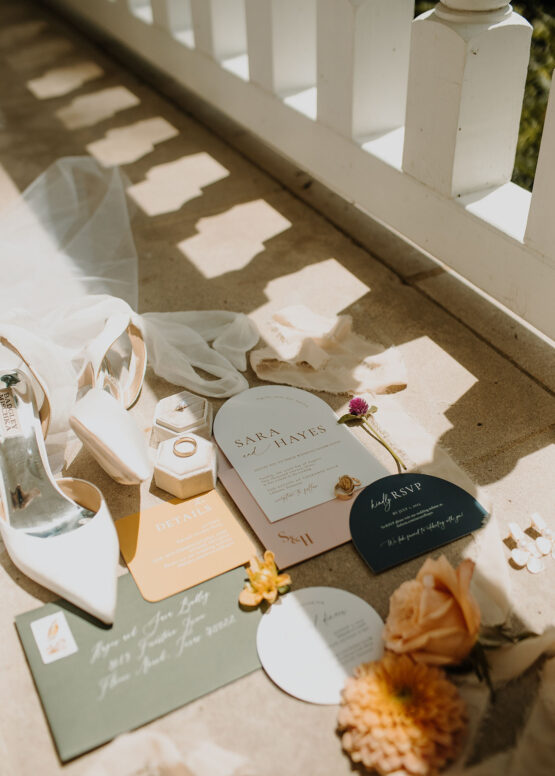 The wedding invitation suite, bridal shoes, and wedding rings laid out in the sunlight on Cherokee Dock's mansion porch