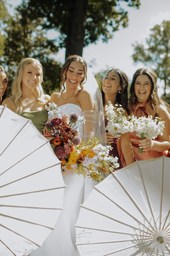 The bride laughs with her bridesmaids as they hold their bouquets and white wedding parasols in front of them
