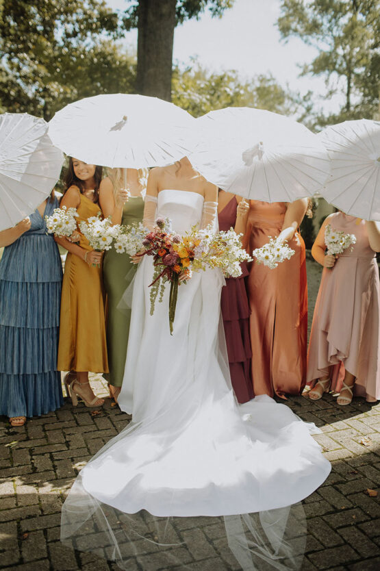 Bridesmaids, each in a different colored dress, hold white bouquets and white wedding parasols over themselves and the bridge, hiding their faces
