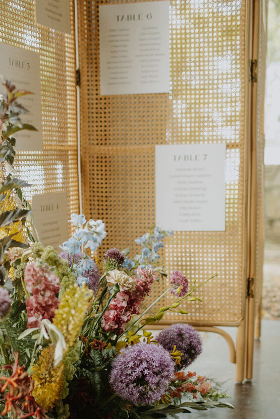 A close-up view of a ground floral arrangement in the foreground with a rattan divider as a seating chart in the background