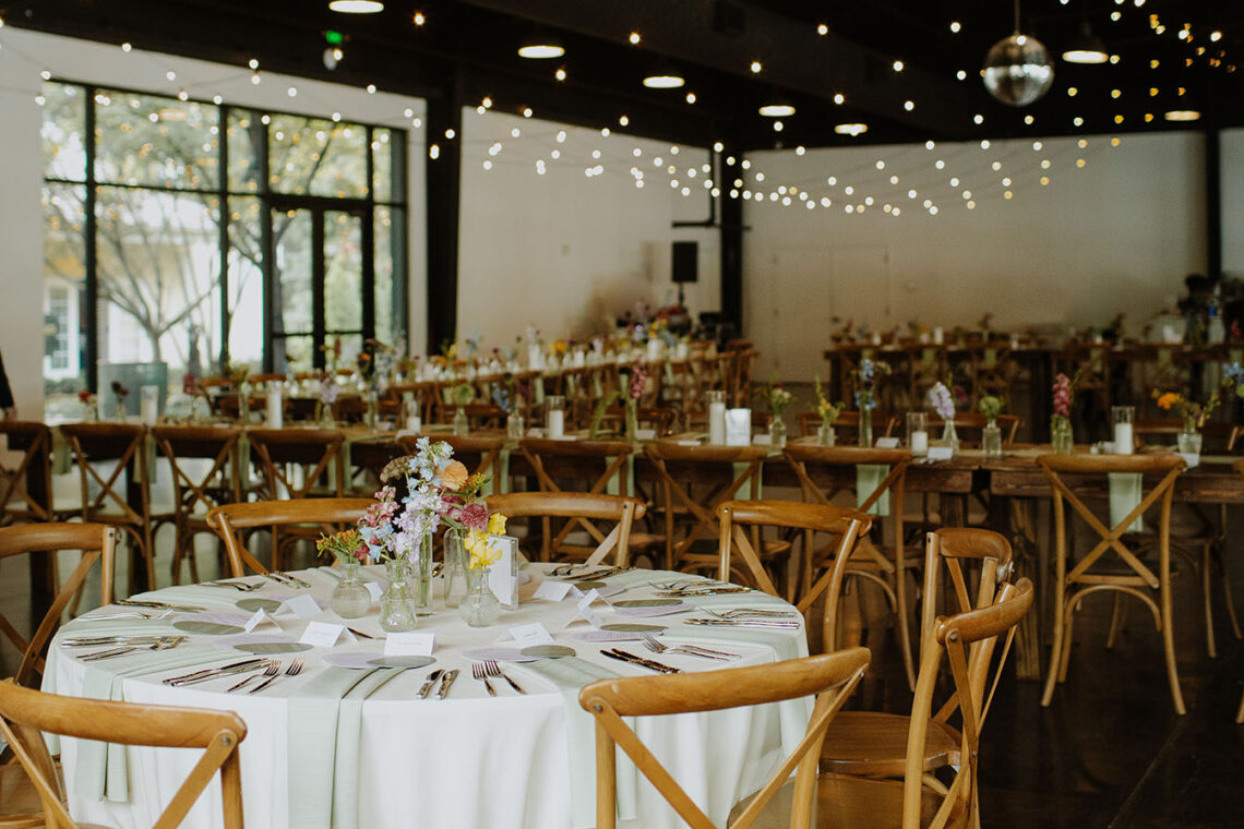 The event center decorated for the reception with light wood chairs, string lights, and a round table with whit linens in the foreground