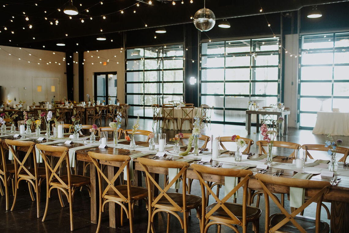 A long farmhouse table in the event center decorated with a white runner, sage green napkins, and small arrangements in bud vases lining the center