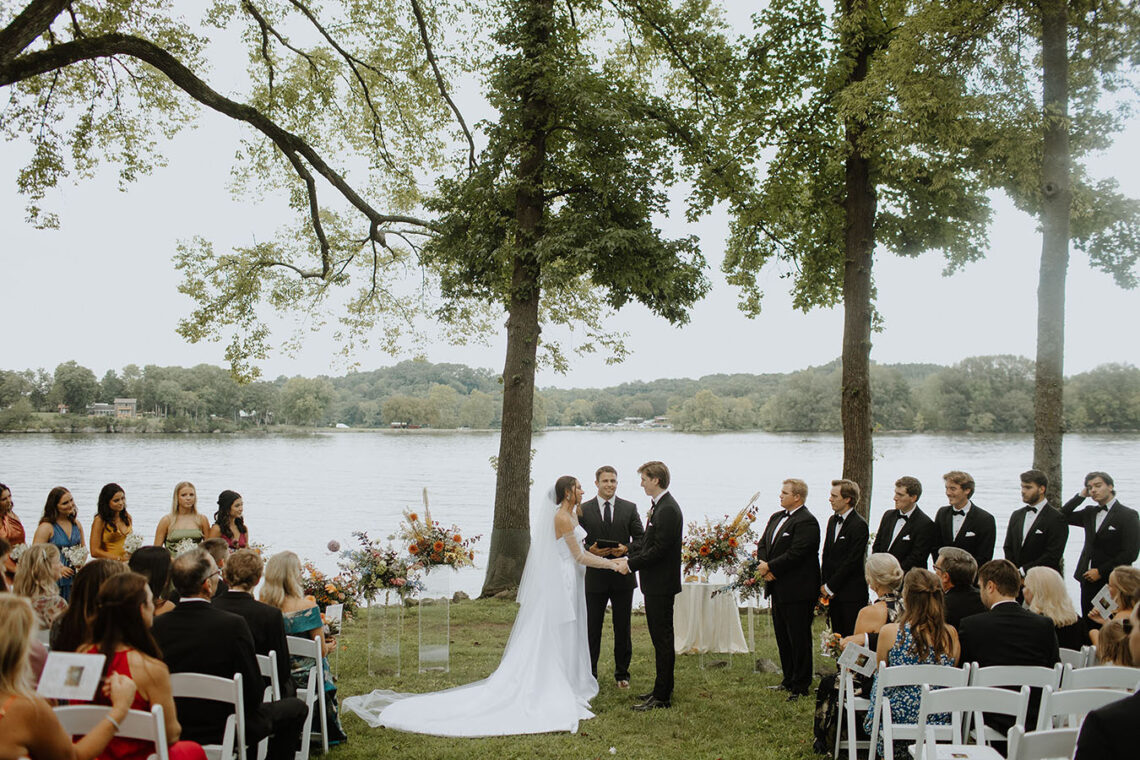 The bride and groom hold hands during their ceremony on the Lakeside Lawn as guests look on