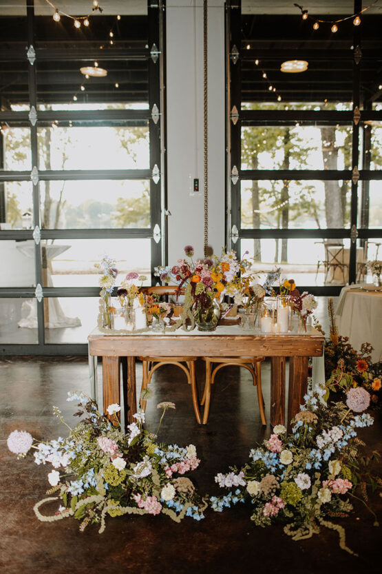 A wooden sweetheart table decorated with vibrant and whimsical floral arrangements with two ground arrangements in the front