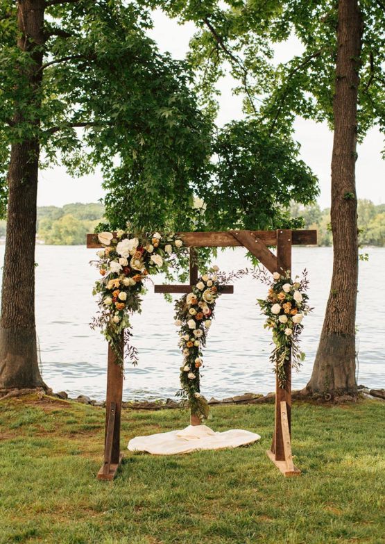 Dark wood ceremony arch adorned with fresh flower arrangements sitting in front of a matching wooden cross
