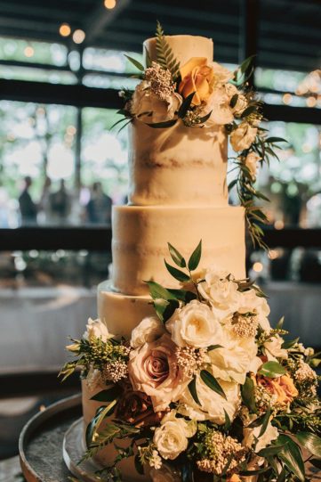 Four-tier wedding cake decorated with fresh roses, mixed greenery, and dainty blooms